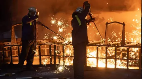 Jon Pountney Two steelworkers in safety jackets, visors and helmets stand in a dark room near orange molten metal, with bright sparks around them.