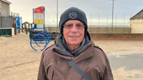 Peter Crome wears a brown jacket and stands on a beach 