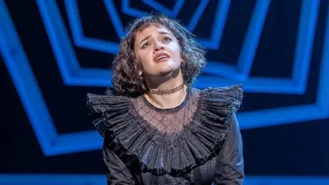 Rachel is wearing a frilly black costume, and a choker around her neck. She has shoulder length black hair and is looking up into the sky.