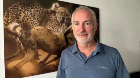 Peter Haygarth stands in front of a large picture of his showing a cheetah and dogs attacking each other. He has short white hair and a neat white beard.