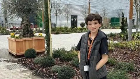 Contributed Tyne, outside, by a building, with buses and trees near him. He has on a lanyard, a black T-shirt, black gilet and black trousers. He is smiling and has his hands in his pockets. He has short dark hair. 