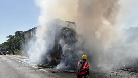 A lorry with clear smoke and fire damage on the road. There is black debris on the road with smoke billowing from the lorry's shell. A small volume of flame can be seen above the front wheel and a firefighter is kneeling down in front of the lorry on the road. 
