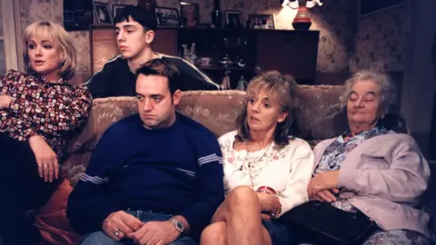 Caroline Aherne, Ralf Little, Craig Cash, Sue Johnston and Liz Smith are sitting on a sofa watching the TV during an episode of The Royle Family.
