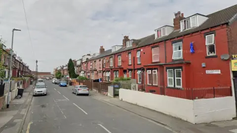 A picture of a residential area. The houses on the right are painted in a coppery red and feature the street sign on the right corner of the first home. Several cars are parked next to the curb on either side of the road.