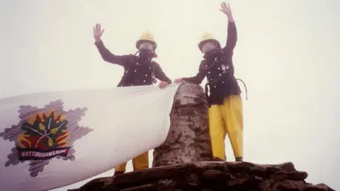 David Needham Two firefighters on a mountain summit wearing full kit and masks, holding a Nottinghamshire Fire Service flag. 