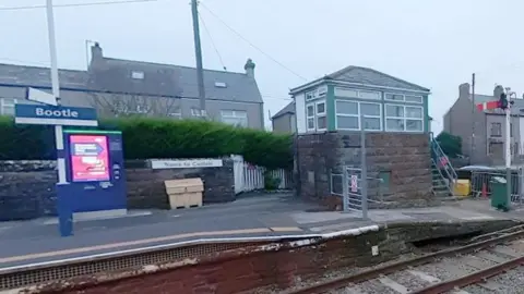 The signal box at Bootle train station in Cumbria. The bottom of it is made out of stone brick and the top is made from wood. It has glass windows and is green and white.