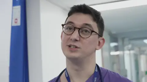 A doctor in purple scrubs has a lanyard around his neck and a stethoscope. He has dark hair and tortoiseshell coloured round glasses.