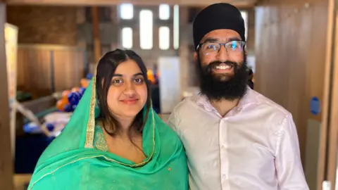 A man and woman stand next to each other smiling. The woman on the left has long dark hair and is wearing a green outfit. The man has a black beard, glasses and is wearing a light pink shirt and Sikh headwear. 