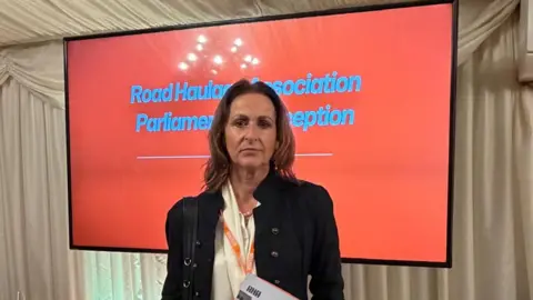 Lesley O'Brien Image shows a lady wearing a black suit. She is standing in front of an orange screen with blue writing on it. The writing says 'Road Haulage Association Parliamentary Reception'. 