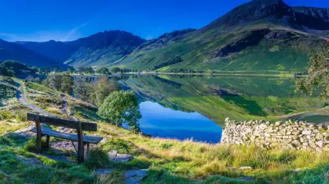 A wooden bench in the foreground overlooks Buttermere. Hills in the distance are reflected in the lake on a sunny day.