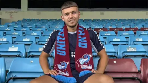 Scunthorpe United FC Jonathan Gjoshe sitting in the stands at Glanford park wearing blue shorts and shirt and a Scunthorpe United scarf