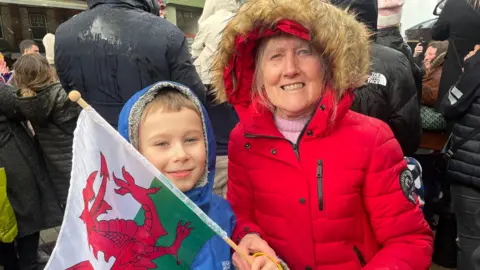 Petricia Thomas is standing to the right wearing a red puff jacket and a hood with fur. He is holding a Welsh flag. On her left, her grandson is Harrison who is wearing a blue raincoat, small golden hair appears under her hood.