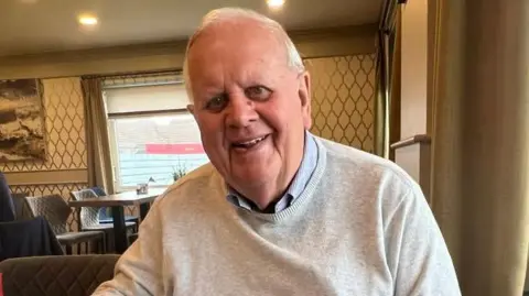 PHIL WOODFORD Photograph of Phil Woodford, who is smiling while sitting in a tearoom. He has grey hair and is wearing a pale grey jumper over a light blue shirt.