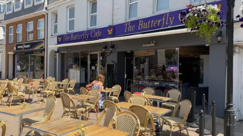Tables outside a cafe called The Butterfly Cafe on a sunny day. The cafe takes up two shop fronts and there is a hanging basket with flowers next to it.