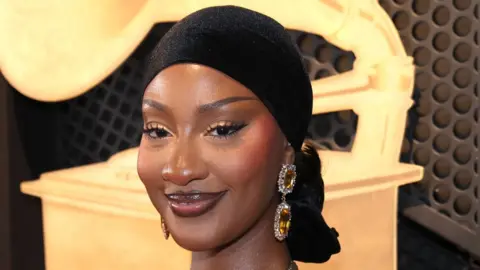 A headshot of Tems wearing a black headscarf smiling and looking at the camera with a large image of golden Grammy behind her.