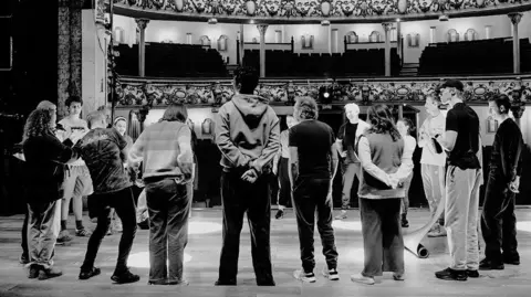 Jon Pountney The entire cast of Our Town are photographed in black and white at Swansea's Grand Theatre. They are stood on stage in a circle. The theatre is empty - it seems to be a rehearsal.