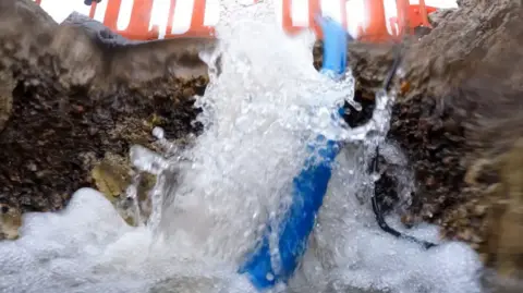 Water is gushing into a hole in a road. The image is close up from inside the hole and looking upward. There is a blue pipe half in and half out of the hole. Above on the road, you can see orange fencing around the hole.