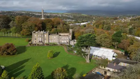 A large historic stone mansion sits in the middle of sweeping green lawns and trees. The estate is a small castle set within a private park. Beyond the grounds are autumn-coloured woodlands and a small town 