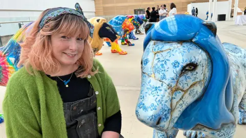 Sam, who has light pink hair, smiles at the camera beside her lion. She wears a blue scarf in her hair, a black top, grey dungarees and a bright green cardigan. Her lion has a bright blue mane, and is covered in a pottery-like blue floral pattern. The surface appears broken, and held together with gold. 