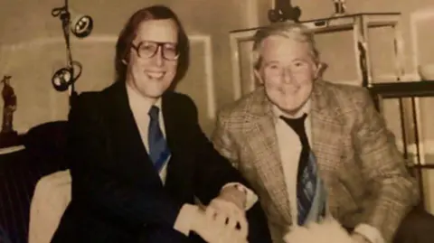 Supplied An old image of Barrie Tomlinson, sitting next to Ernie Wise. Barrie has glasses on and a dark suit, white shirt and tie and Ernie a checked suit jacket and tie, with short grey hair. They are both smiling and looking straight at the camera.