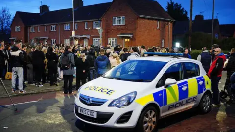 PA Media Members of the public observe a minute's silence during a vigil near to the scene in south Bristol where two teenage boys, aged 15 and 16, died after a stabbing attack. A police car is in the foreground. 