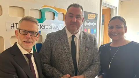 Stagecoach East Three people standing in a row looking towards the camera. On the left is Darren Roe, in a dark suit, white shirt and maroon tie. He has white hair and close-cropped white beard and is wearing rectangular dark framed glasses. Next to him is Paul McCarthy, in a black and white speckled jacket, white shirt and black tie. He is clean-shaven and has short dark spikey hair. Next to him is Hema Russell, who is waring a dark dress or top and a long bar on a long chain. She has pulled back dark hair, tucked behind her ears.