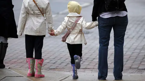Tom Williams/Getty The picture shows a child in the centre of the frame. She is wearing wellingtons and a light-coloured mac. She holds hands with an adult to her right and another child to her left.
