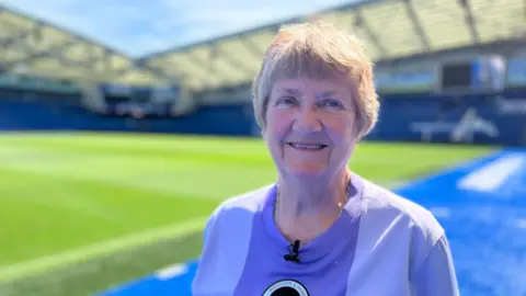 A woman with short grey hair is wearing a purple football shirt and is standing on the side of the football pitch in Brighton & Hove Albion's football stadium.