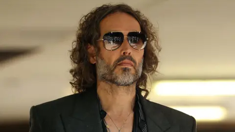 Reuters A headshot of Russell Brand who wears a suit and sunglasses.