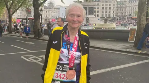 Sue Nicholls is standing on a road in London, wearing a running outfit and a medal. She is smiling.