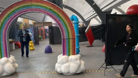 A man walks under a rainbow prop in a largely empty warehouse with a blue pipe and other small colourful props scattered about the floor.