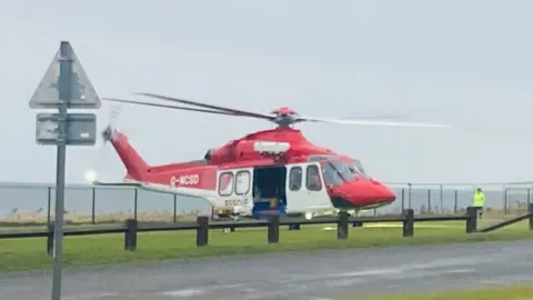 A red and white helicopter on grass in front of a fence and the North Sea. A person in a green hi-vis jacket is stood the the side of the helicopter.