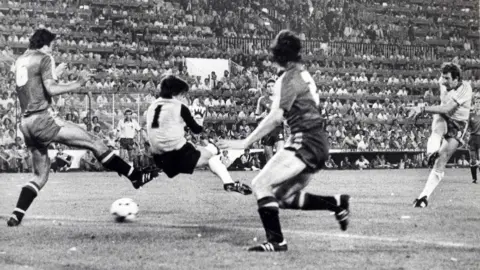 Getty Images A black and white image of Gerry Armstrong scoring against Spain in 1982. Armstrong is on the far right of the image having taken a shot, he is wearing a white football shirt, dark shorts and white socks. The ball is seen going under the goalkeeper who has black hair and is wearing a light jersey with "1" on the back and dark shorts and white socks. Two other Spanish players are near hmi, both in dark kits. The crowd can be seen watching on from the stands.
