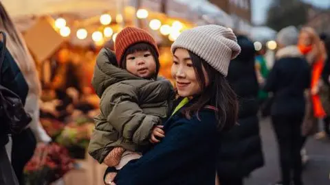 A mother holds a toddler as they stand in a Christmas market. The mother wears a beige beanie and navy coat, and the child wears an orange beanie and a green puffa jacket. 