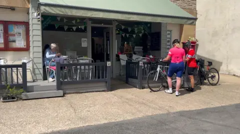 Two cyclists wearing pink and red lycra are standing next to their bicycles outside Thorncombe Village Shop. The shop has a wooden patio area outside its front door with a woman drinking a cup of coffee at a metal table set up.