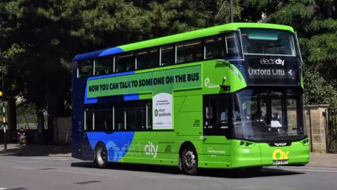 A green electric Oxford City bus on a street. The words 'Now you can talk to someone on the bus' are printed on the side of the vehicle. It is a sunny day.