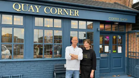 Paul Marriner and Joanne Horner stand in front of their coffee shop. The front is blue. A gold plated sign reads: "QUAY CORNER". Mr Marriner is crossing his arms.