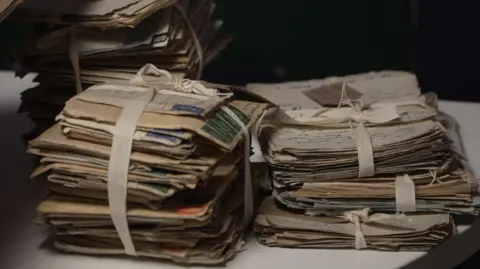 Three stacks of hand-written letters resting on a table. They are held in their piles by white bows, which are tied around them.