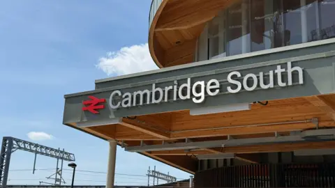 A train station's entrance has a sign above it that says Cambridge South. There are overhead cables on the left where a train line runs. 