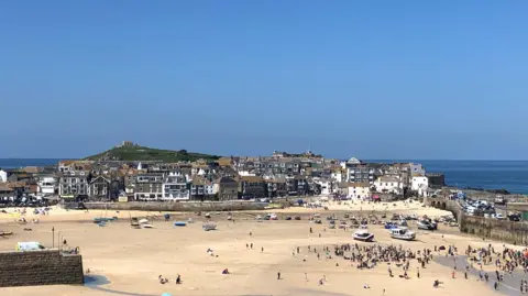 A wide view of St Ives harbour and island with a clear blue sky and the tide out  -exposing golden sand with large clusters of people and boats scattered around. There is also a glimpse of blue sea in the background