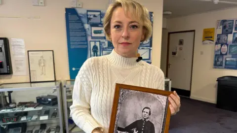 A blonde woman in a white jumper holds a black and white photograph of a man in a police uniform. She is standing in front of a museum display. 