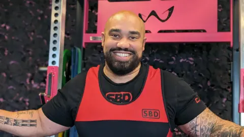 Ram Patten. He is bald and has black facial hair. He is wearing black and red gym clothing and is sat by some gym equipment. He has his arms out, showing his tattoos. Ram is looking directly at the camera and smiling 