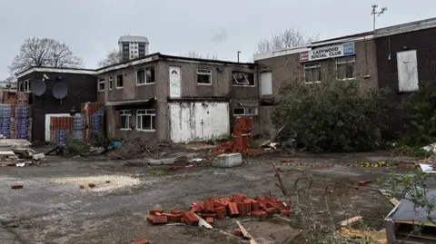 A two-storey building with parts demolished seen from an overgrown yard with piles of pallets and bricks. It is daytime on a grey day. High rise flats and the tips of trees can be seen in the background. A sign on the building says Ladywood Social Club.