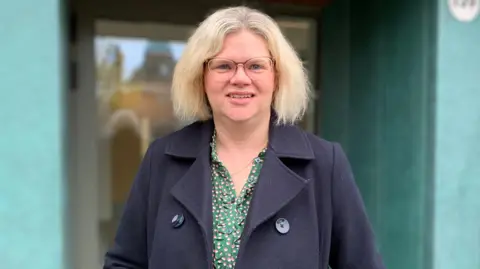 A blonde haired women with glasses wearing a dark coat with a green patterned blouse stands in front of a green doorway