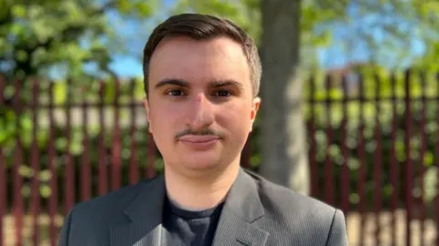 Head and shoulder image of Elliot Tong. He has short dark hair, a moustache and is wearing a grey suit jacket and dark top.