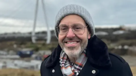 Leo Pearlman wearing a grey beanie hat and a blue overcoat, and gold thin-rimmed glasses, stands in front of a construction site. This is set to become a TV and film studio in Sunderland.