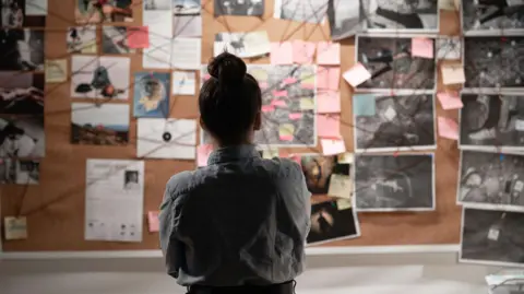 A female police officer wearing a pale blue collared shirt, with her dark hair tied up in a high bun. She is facing away from the camera, looking at a large notice board on the wall which lays out an organised crime investigation. There are lots of post-it notes and images pinned to the wall, all connected with red string in different directions.