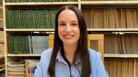 Claire has long brown hair, she is wearing a blue and whit striped shirt. Behind her are wooden book shelves filled with books.