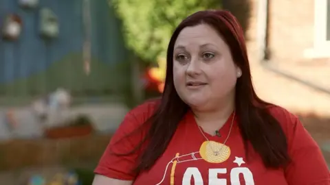 Ellie has long dark-red hair and wears a red T-shirt with the AFO logo on it.  She is sitting outside for her TV interview.