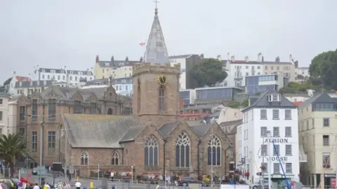 A tradition church, with a clock. The tower on top of the church is made of glass and has a cross. There are houses around the church and a road and cars. 